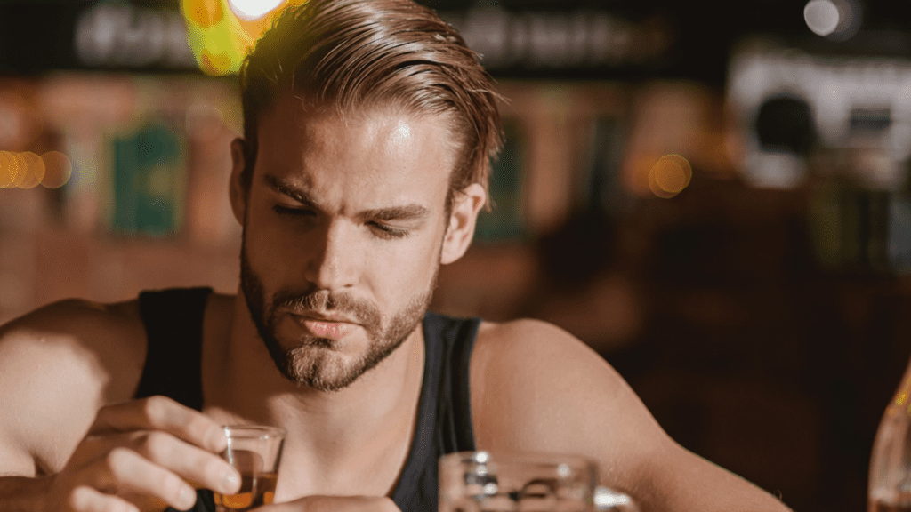 A gay person sitting on a table with a glass of alcohol thinking about getting drinking addiction help.