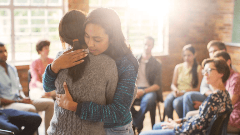 Two people hugging at an AA meeting with others sitting in a circle, symbolizing support and healing