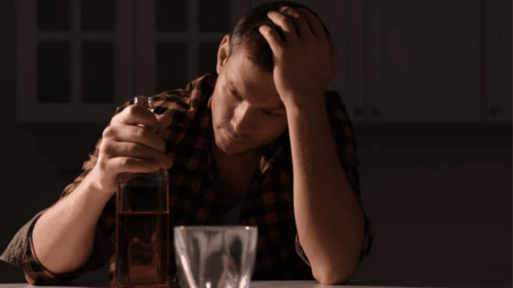 Gay man leaning on his hand, looking distraught with a bottle of alcohol and an empty cup, symbolizing struggles with alcoholism