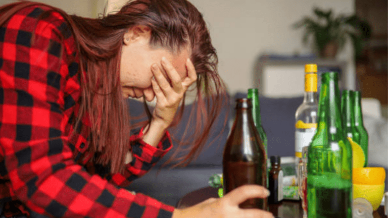 A queer person holding a bottle of beer on a table, showing distress and the struggles from alcoholism symptoms.