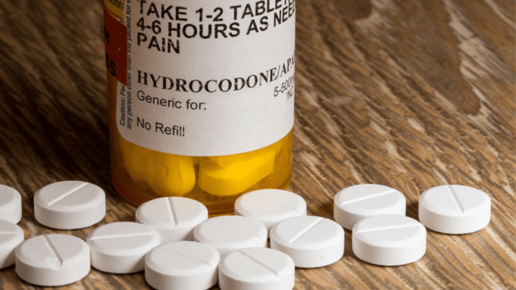 Prescription bottle labeled Hydrocodone with white tablets scattered on a wooden surface as example of pain medication.