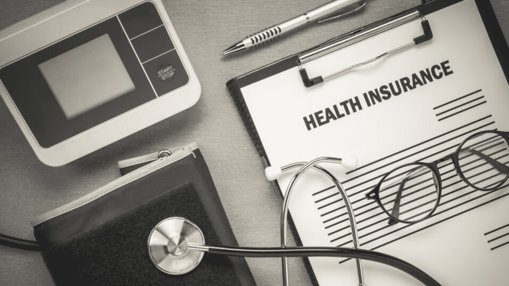 Clipboard with a health insurance form, pen, glasses, stethoscope, and blood pressure cuff on a table.