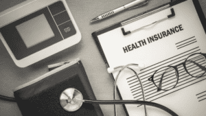 Clipboard with a health insurance form, pen, glasses, stethoscope, and blood pressure cuff on a table.