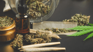 Close-up of cannabis buds, rolled joints, a dropper bottle, and a grinder on a table, showing marijuana use and preparation.