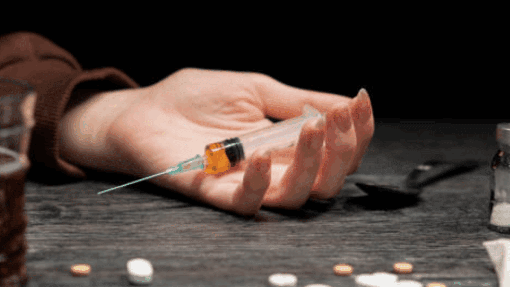A hand of a person high on fentanyl while holding a syringe with fentanyl content on a table.