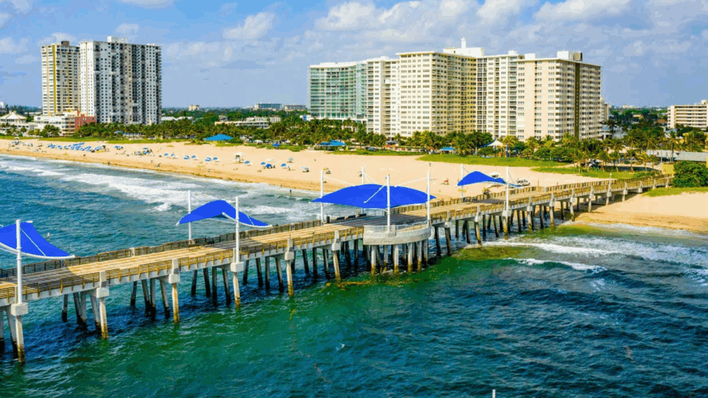 A scenic view of Pompano Beach, Florida, with a pier extending over the ocean and high-rise buildings in the background.