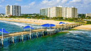 A scenic view of Pompano Beach, Florida, with a pier extending over the ocean and high-rise buildings in the background.