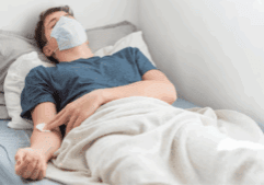 A masked gay man lying on a hospital bed.