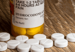 Prescription bottle labeled Hydrocodone with white tablets scattered on a wooden surface as example of pain medication.