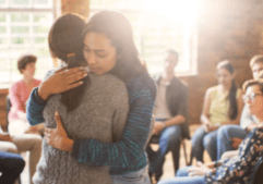 Two people hugging at an AA meeting with others sitting in a circle, symbolizing support and healing