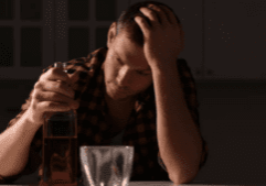 Gay man leaning on his hand, looking distraught with a bottle of alcohol and an empty cup, symbolizing struggles with alcoholism