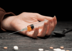 A hand of a person high on fentanyl while holding a syringe with fentanyl content on a table.